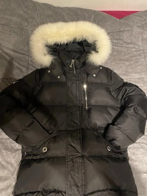 Svart dunjacka från Moose Knuckles - Snygg svart dunjacka från Moose Knuckles med stor huva och vit fuskpäls. Jackan har dragkedja, tryckknappar och flera fickor, inklusive en bröstficka med dragkedja. Perfekt för kalla vinterdagar och riktigt varm.