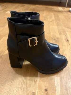 Svarta stövletter med klack och spänne - Snygga svarta stövletter med bred klack och markerad platåsula. Dekorativt spänne i silver på sidan och dragkedja på insidan för enkel på- och avtagning. Perfekta för dig som vill ha en edgy och trendig look. Stl. 39! Enbart testade inomhus! 