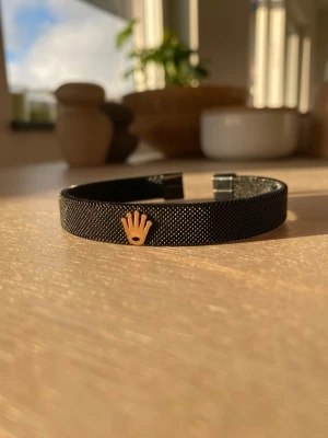 Svart armband med Rollex-logga - Stilrent svart armband i metall med meshstruktur och en guldfärgad Rollex-krona framtill. Öppen design som gör det enkelt att ta på och av. Perfekt accessoar för dig som gillar exklusiva detaljer och vill ha något som sticker ut.