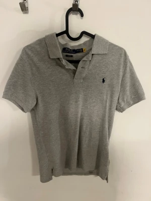 Grå pikétröja från Polo Ralph Lauren - Klassisk grå pikétröja från Polo Ralph Lauren med korta ärmar och krage. Tröjan har den ikoniska broderade loggan på bröstet och är tillverkad i mjuk bomull. Perfekt till jeans eller shorts för en avslappnad look.