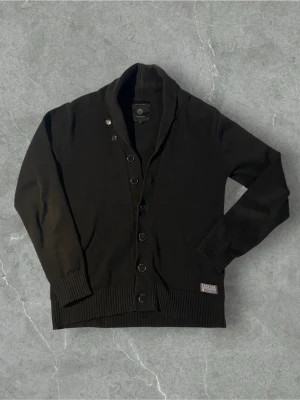 Svart kofta från Premium by Jack & Jones | L - Stilren svart kofta med knappar framtill från Premium by Jack & Jones. Koftan har ribbade muddar vid ärmslut och nederkant samt en liten tygpatch nertill. Perfekt för lager på lager och enkel att matcha med olika outfits.