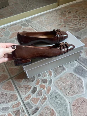 Bruna skinnimitation ballerinaskor från Tamaris - Snygga bruna ballerinaskor i lack från Tamaris med låg klack och dekorativa dubbla spännen framtill. Skorna har en klassisk rund tå och är tillverkade i glansigt material som ger en elegant touch. Perfekta för dig som gillar stilrena detaljer. Använda endast vid ett tillfälle 🌸