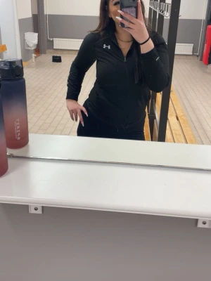 Svart träningsjacka från Under Armour - Säljer en svart träningsjacka från Under Armour med dragkedja framtill och hög krage. Jackan har en slim fit passform och är tillverkad i ett stretchigt funktionsmaterial som passar perfekt till gymmet eller löprundan.