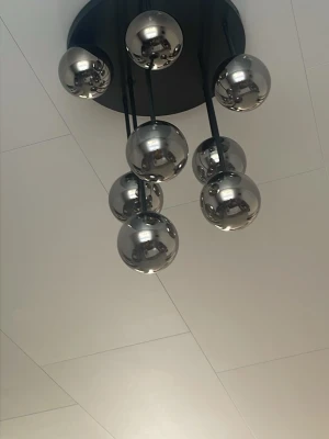 Modern taklampa med kromade klot - Säljer en modern taklampa med sju spegelblanka, kromfärgade klot i metall. Lampan har en rund svart takplatta och svarta stänger som håller upp kloten i olika höjder. Perfekt för dig som gillar stilren och futuristisk design.