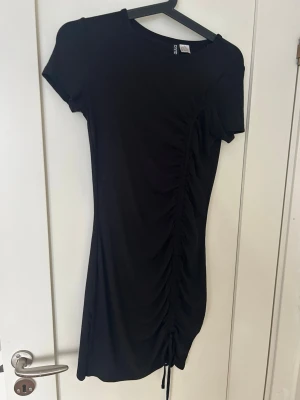 Svart kortklänning med snörning H&M  - En svart kortklänning från H&M med snörningsdetalj längs sidan som ger en snygg rynkeffekt. Klänningen har rund halsringning och korta ärmar. Tillverkad i mjukt stretchigt material som sitter bekvämt och följer kroppen.