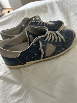 Blåa sneakers från Philippe Model - Snygga blåa sneakers från Philippe Model med grå mockadetaljer och vita snören. Skorna har en klassisk låg profil och en vit sula som ger en clean look. Perfekta för dig som gillar att sticka ut!