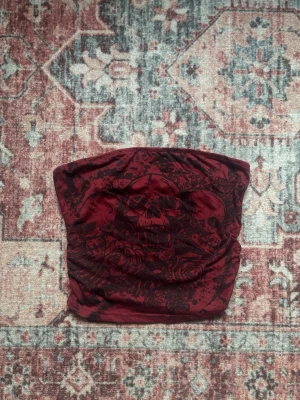 Vinröd bandeau tubtopp med dödskalle - Snygg vinröd bandeau tubtopp med tryck av dödskalle och rosor framtill. Toppen är axelbandslös och har en tight passform som sitter på plats. Är croppad.