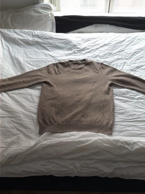 Beige cashmeretröja från Piacenza - Mjuk och stilren beige stickad tröja från lyx märket Piacenza i 100% cashmere. Tröjan har rund halsringning, långa ärmar och ribbade muddar vid ärmslut och nederkant. Perfekt för dig som gillar klassisk och enkel stil. Har ett reparerat hål på magen som knappt är märkbart och ett på en av armuddarna som inte heller är märkbart. 