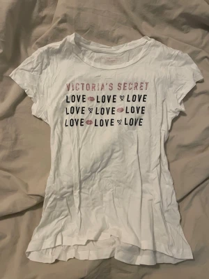 Vit t-shirt Victoria's Secret - Vit t-shirt från Victoria's Secret med tryck i rosa och svart text där det står 'LOVE' och 'VICTORIA'S SECRET' samt små läppar och hjärtan. Klassisk rund hals och korta ärmar. Perfekt för en avslappnad och trendig look.
