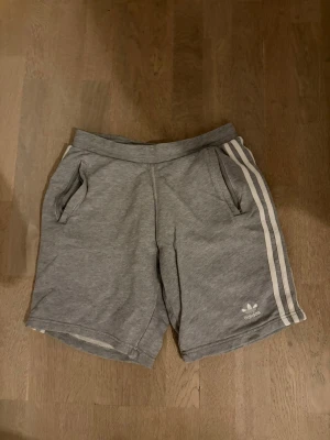 Grå Adidas shorts med vita ränder - Klassiska grå shorts från Adidas med vita signaturränder längs sidorna och Adidas-logga på vänster ben. Mjuk resår i midjan och två fickor framtill. Perfekta för en avslappnad och sportig stil.