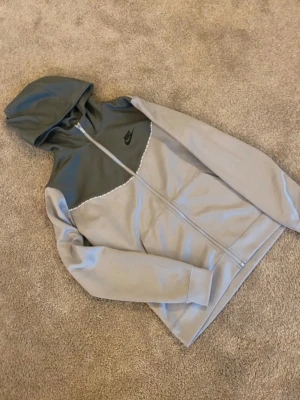 Nike hoodie i grått och mörkgrått - Snygg hoodie från Nike i två nyanser av grått med dragkedja framtill och huva. Den har en svart Nike-logga på bröstet och är tillverkad i ett mjukt och bekvämt material, perfekt för en avslappnad stil.