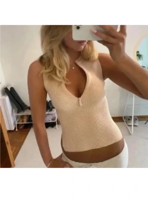 Beige ribbad ärmlös topp - Snygg beige ribbad topp med v-ringning och liten dragkedja framtill. Toppen är ärmlös och har en croppad passform som framhäver midjan. Perfekt för dig som gillar en enkel men trendig stil.