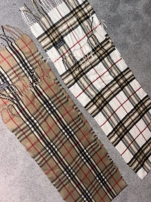 Burberrys rutiga halsdukar i ull - Två klassiska halsdukar från Burberrys i 100% lammull. Ena halsduken är beige med svart, vitt och rött rutmönster, den andra är vit med samma rutiga design. Båda har fransar i ändarna och ikoniska Burberrys-märket. Perfekt accessoar för en tidlös look.