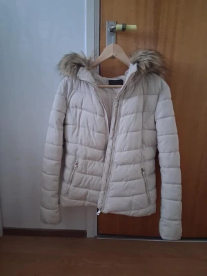 Beige pufferjacka med fuskpäls - Snygg beige pufferjacka med avtagbar huva och fuskpälsdetalj. Jackan har två dragkedjefickor framtill och quiltad design som ger en modern look. Perfekt för kalla vinterdagar och håller dig varm och stilren.