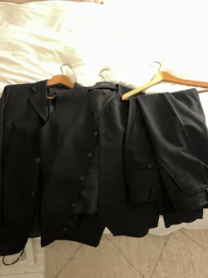 Svart kostym med väst till - Säljer en klassisk svart kostym med matchande väst som är skräddarsydd från Thailand. Kostymen har tvåknäppt kavaj, raka byxor och enkel design utan synliga detaljer. Perfekt för dig som vill ha en stilren och tidlös look. Materialet är troligtvis polyesterblandning. Dåligt ljus på bilderna. Måttet på personen som burit kavajen är 178cm och väger 75kg. Fler bilder finns. 