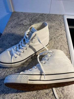 Vita höga canvas sneakers - Säljer ett par klassiska vita höga sneakers i canvas med vita snören och gummisula. Skorna har metallhål för snörningen och svart rand längs sulan. Perfekta för en clean och stilren look.