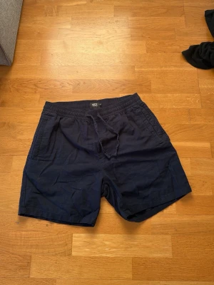 Mörkblå shorts från 157 med snörning - Säljer ett par mörkblå shorts från 157 i bomull. De har elastisk midja med snörning och två sidofickor. Perfekta för varma dagar och enkel stil.