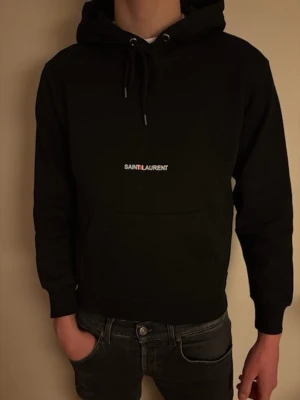 Saint Laurent hoodie svart XS - Saint Laurent hoodie – XS ⚜️  Stilren Saint Laurent hoodie i storlek XS. Klassisk svart färg med diskret logga framtill. Hoodien har blivit något mindre i tvätten, men sitter fortfarande snyggt. Inga defekter i övrigt och i bra skick.  ⚜️ Märke: Saint Laurent ⚜️ Storlek: XS ⚜️ Färg: Svart ⚜️ Skick: Bra skick, inga defekter ⚜️ Info: Något krympt i tvätten ⚜️ Modellen på bilden är 177 cm och väger 57 kg  Mått (platt mätta): ⚜️ Midja till axel: 56 cm ⚜️ Axel till axel: 45 cm  Redo att skickas 📦⚜️ 