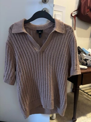 Beige ribbad topp med krage H&M - Säljer en beige ribbad topp från H&M med v-ringning och krage. Toppen har korta ärmar och är gjord i ett mjukt stickat material. Perfekt för dig som gillar en avslappnad men ändå stilren look.
