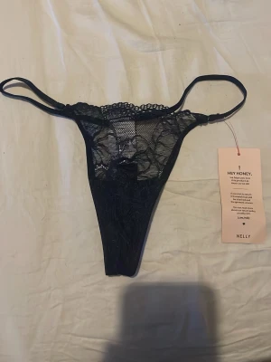 Mörkblå spetsstring från Nelly - Snygg mörkblå stringtrosa från Nelly i modellen Delicate Bow Thong. Trosan har tunna dubbla sidoband och är gjord i transparent spets med blommigt mönster och liten rosett framtill. Perfekt för dig som gillar feminina detaljer och en trendig look.