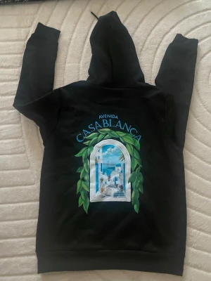 Svart hoodie Casablanca Avenida - Svart hoodie från Casablanca med stor färgglad grafik på ryggen som föreställer ett fönster med utsikt mot havet och gröna blad runtom. På bröstet finns en mindre version av samma motiv och texten 'Avenida Casablanca'. Klassisk känguruficka och huva.