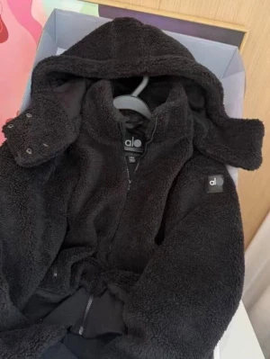 Svart teddyjacka med huva från Alo - Mysig svart teddyjacka från Alo med dragkedja framtill och stor huva. Jackan har en fluffig, mjuk yta och ribbade muddar vid ärmsluten. Märkets logga syns på bröstet och insidan av kragen. Perfekt för kalla dagar och riktigt skön att bära.