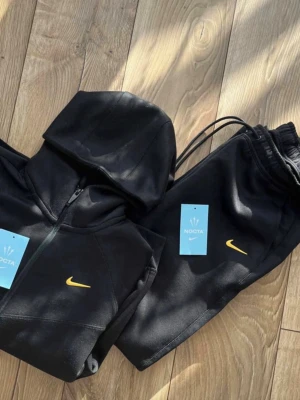 Svarta NOCTA x Nike mjukisbyxor - Säljer ett par svarta mjukisbyxor från Nike och NOCTA-kollektionen. Byxorna har gul NOCTA-logga på benet och en liten gul Nike Swoosh vid fickan. Utrustade med dragkedjefickor och snörning i midjan. Perfekta för en chill och sportig look.