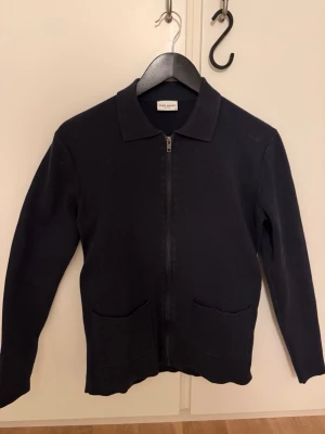 John Henric Ziptröja - Klassisk svart ziptröja från John Henric med krage och två framfickor. Tröjan har lång ärm och rak passform, perfekt att slänga över en t-shirt. Passar s/xs