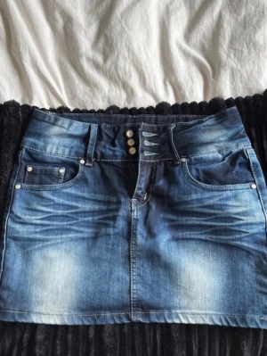 Blå jeanskjol med knappar - Snygg blå jeanskjol i kort modell med slitningar och dekorativa knappar framtill. Kjolen har klassiska fickor både fram och bak samt bälteshällor. Perfekt för dig som gillar en avslappnad och trendig stil.