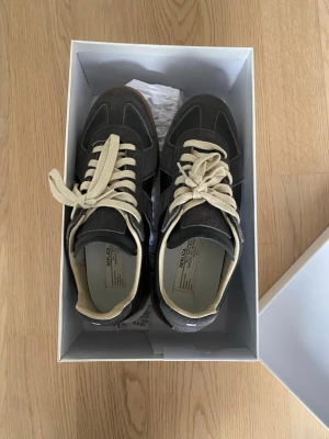 Svarta sneakers från Replica Maison Margiela - Snygga svarta sneakers från Replica Maison Margiela med beigea detaljer och vita snören. Skorna har en klassisk låg siluett och är tillverkade i mocka och läder för en stilren look. Perfekta för dig som gillar minimalistisk och trendig design.