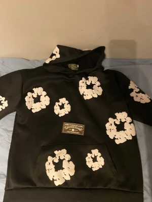 Svart set med vita blommor från Denim Tears - Säljer ett svart set från Denim Tears med matchande hoodie och byxor. Setet har stora vita blommönster tryckta över hela plaggen och en ficka framtill på hoodien. Materialet ser ut att vara mjukt och bekvämt, perfekt för en avslappnad stil.