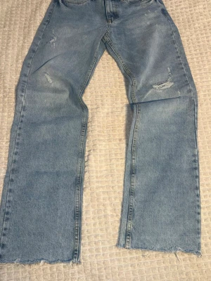 Ljusblå raka jeans från Guess - Säljer ett par ljusblå jeans från Guess med raka ben och råa, fransiga benslut. Jeansen har slitna detaljer och klassisk femficksdesign. Materialet är denim i bomull och bakfickan har Guess-loggan. Perfekta för en avslappnad och trendig look.