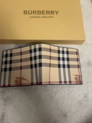 Burberry plånbok i beige rutigt mönster - Säljer en klassisk Burberry plånbok i beige med det ikoniska rutiga mönstret i svart, vitt och rött. Plånboken är tillverkad i canvas med detaljer i läder och har Burberrys logga tryckt på insidan. Perfekt för dig som gillar stilrena accessoarer.