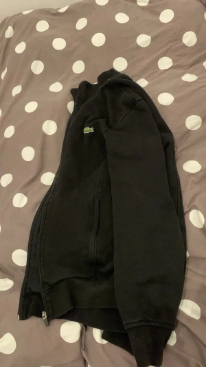 Svart Tröja från Lacoste - Säljer en svart Tröja från Lacoste med dragkedja framtill och det klassiska gröna krokodilmärket på bröstet. Hoodien har långa ärmar, ribbade muddar och två fickor på sidorna. Storleken är en lite liten medium! Den sitter alltså rätt tight 