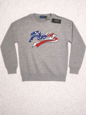 Grå sweatshirt från Polo Ralph Lauren - Snygg grå sweatshirt från Polo Ralph Lauren med stort tryck på bröstet där det står 'Polo' i blått, vitt och rött med stjärnmönster. Tröjan har rund halsringning, ribbade muddar och är perfekt för en avslappnad streetwear-look. Skriv vid frågor och funderingar. Pris kan diskuteras!
