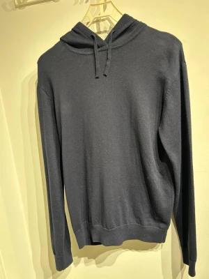 Mörkgrå hoodie med huva - En stilren mörkgrå hoodie med huva och dragsnören. Tillverkad i ett mjukt, stickat material som ger en avslappnad och bekväm känsla. Hoodien har långa ärmar och ribbade muddar vid ärmslut och nederkant. Perfekt för en enkel och clean look.