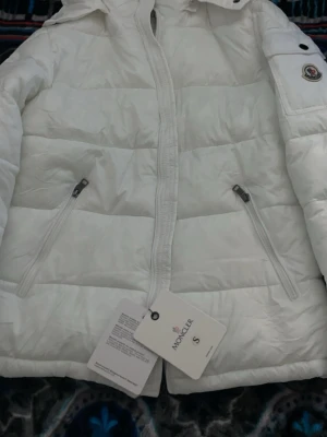 Vit pufferjacka från Moncler S - Snygg vit pufferjacka från Moncler med två dragkedjeförsedda fickor framtill och klassisk Moncler-logga på ärmen. Jackan har quiltad design och hög krage. Materialet är glansigt och ger en exklusiv känsla. Perfekt för kalla dagar när du vill hålla stilen.