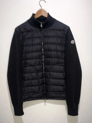 Svart cardiganjacka från Moncler - Snygg svart cardiganjacka från Moncler med quiltad front och stickade ärmar. Dragkedja framtill och klassisk Moncler-logga på ärmen. Perfekt för lager-på-lager och streetwear-vibe. Materialmix av dun och bomull för både värme och stil.