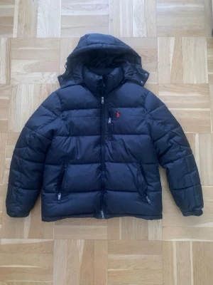 Svart dunjacka från Polo Ralph Lauren - Snygg svart dunjacka från Polo Ralph Lauren med röd broderad logga på bröstet. Jackan har huva, två dragkedjeförsedda fickor och quiltad design. Perfekt för kalla vinterdagar och riktigt stilren look.