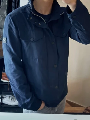 J.Lindeberg field jacket  - Snygg marinblå Field jacket från J.Lindeberg i storlek M med hög krage, bröstfickor med lock och tryckknappar samt diskret logga på ärmen. Jackan har dragkedja och knappslå framtill och är tillverkad i ett lätt, slitstarkt material. Perfekt för dig som gillar stilren och funktionell design.