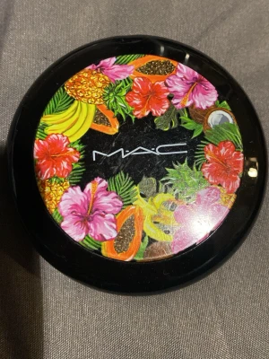 MAC bronzer med tropiskt lock - Snygg bronzer från MAC i en rund svart dosa med spegel och ett färgglatt tropiskt motiv på locket. Pudret har en varm, matt brun nyans som ger huden en solkysst look. Perfekt för att framhäva ansiktskonturer och skapa glow.