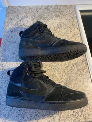 Nike svarta skor - Säljer ett par helsvarta Nike high top sneakers med klassisk swoosh på sidan. Skorna har en kombination av mocka och textil, samt snörning hela vägen upp. Insidan är fodrad med mjukt material för extra komfort. Perfekta för dig som gillar streetwear och sportig stil. Jätte sköna, bekvema o fina skor. 