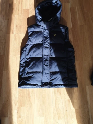 Svart dunväst från Lyle & Scott - Snygg mörkblå dunväst från Lyle & Scott med huva och gul logga på bröstet. Västen har quiltad design och dragkedja framtill. Perfekt för lager på lager och håller dig varm under kyliga dagar. Knappen är lite trasig längst ner men funkar och är bra ändå.. Kolla sista bild