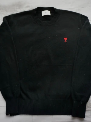 Svart stickad | Ami Paris - Svart stickad tröja från Ami Paris med rund halsringning och ribbade muddar. På bröstet finns Ami:s ikoniska röda hjärtemblem. Tröjan har en klassisk passform och är perfekt för lager-på-lager. Materialet ser ut att vara mjukt och av hög kvalitet.