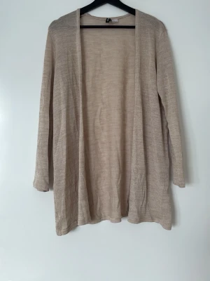 Beige lång öppen kofta från H&M - Lång, öppen kofta i tunn beige trikå från  H&M. Koftan har långa ärmar och en avslappnad passform utan knappar eller dragkedja. Perfekt att slänga över ett linne för en enkel och stilren look.