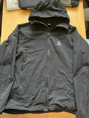 Svart vindjacka från Haglöfs med huva - Snygg svart vindjacka från Haglöfs med hel dragkedja framtill, två sidofickor med dragkedja och justerbar huva. Jackan har minimalistisk design och diskret Haglöfs-logga på bröstet. Perfekt för blåsiga dagar och enkel att packa ner i väskan.