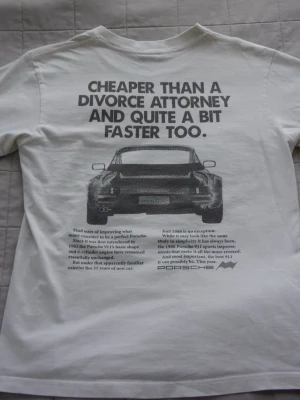 Vit Porsche t-shirt med tryck - Vit t-shirt från Porsche med stort svart tryck på ryggen som visar en klassisk Porsche 911 och texten 'Cheaper than a divorce attorney and quite a bit faster too.' T-shirten har rund hals och korta ärmar. Perfekt för dig som gillar bilar och retrostil.
