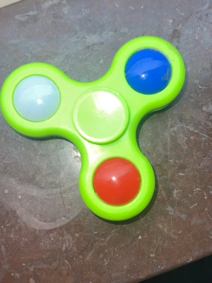Grön fidget spinner med färgknappar - En grön 💚fidget spinner i plast med tre stora knappar i rött❤️, blått💙 och vit🤍t. Spinnern har rundade kanter och är lätt att snurra mellan fingrarna. Perfekt för att hålla händerna sysselsatta eller som stressleksak. Den lyser också när man snurrar på den
