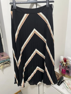 Plisserad svart långkjol med ränder - Snygg plisserad långkjol från Next i svart med breda, diagonala ränder i beige och vitt. Kjolen har ett flowy fall och elastisk midja för skön passform. Perfekt att styla med en enkel topp och sneakers.