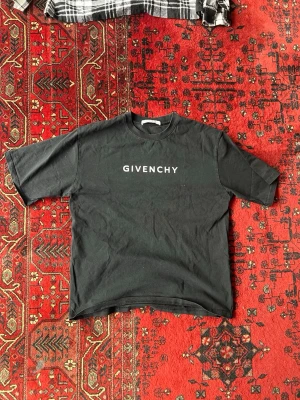 Svart Givenchy t-shirt med tryck - Säljer en svart t-shirt från Givenchy med vit logga framtill och ett stort kors med texten 'Paris Givenchy' i vitt och rött på ryggen. T-shirten har rund hals och korta ärmar. Materialet är mjuk bomull och passformen är avslappnad.